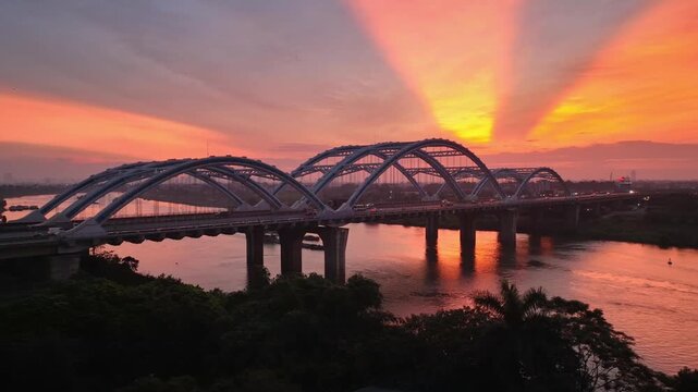 Aerial view of Dong Tru Bridge crossing the Red River at sunset, with dramatic orange skies, shimmering water, and the Hanoi skyline in the distance.