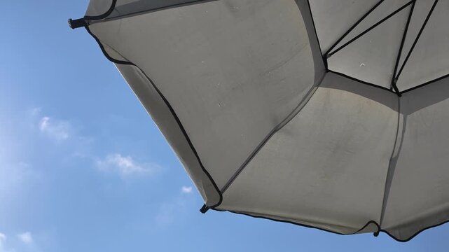 White patio umbrella spinning slowly in the gentle breeze against a bright blue summer sky with wispy clouds, creating a relaxing and peaceful holiday atmosphere