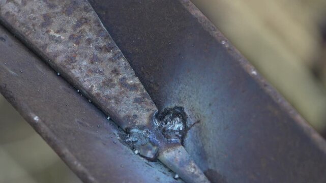 Hardened slag chips away as a fabricator cleans freshly welded steel joints in a messy workshop. Extreme close-up macro shot.