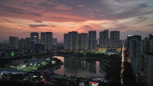 Aerial view of Hanoi's illuminated high-rise buildings, calm urban lake, winding roads, and traffic along Le Van Luong Street at sunset.