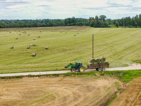 Aerial view of a green tractor pulling a trailer loaded with round hay bales through a vast agricultural landscape in Talsi Municipality, Latvia.
