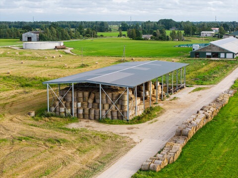 Aerial view of a farm featuring a large open-sided shed stacked with round hay bales, with additional bales lined up along a dirt road in the rural landscape of Virpe, Ventspils novads, Latvia.