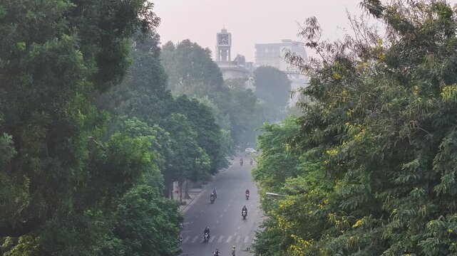 A tree-lined Dinh Tien Hoang Street near Hoan Kiem Lake in Hanoi, with motorbikes traveling through lush greenery beneath a hazy urban skyline.