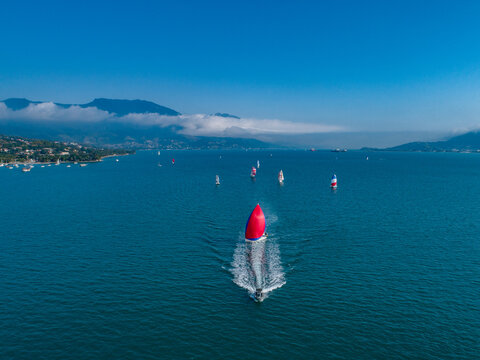 Aerial view of a sailboat with a vibrant red spinnaker sailing across the blue water with other boats in the distance near the coast of Ilhabela, Sao Paulo, Brazil.