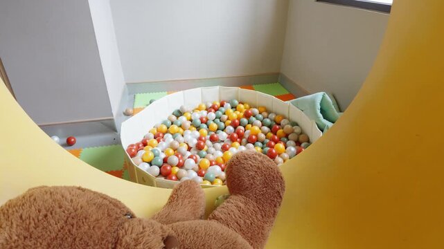Teddy bear sitting on a yellow indoor slide overlooking a colorful ball pit in a children playroom.