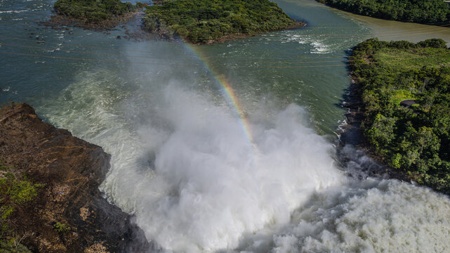Aerial view of Furnas Hydroelectric Plant with water rushing through the spillway creating a rainbow over the river, surrounded by lush vegetation in Rio Grande, Minas Gerais, Brazil.