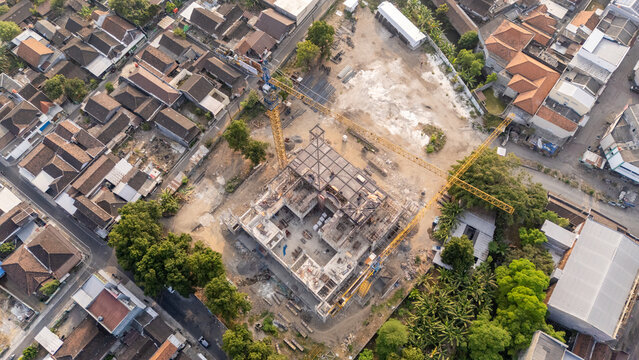Aerial view of a construction site with a yellow tower crane, steel framework, and building materials surrounded by residential houses and trees in Surakarta, Central Java, Indonesia.