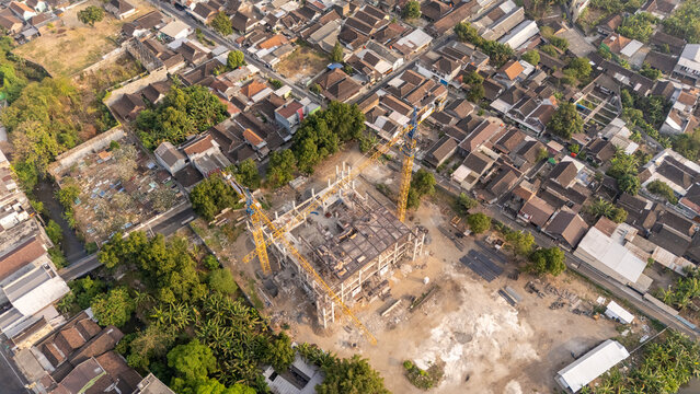 Aerial view of a construction site featuring a yellow crane amidst a dense residential neighborhood with rooftops and lush trees in Surakarta, Central Java, Indonesia.