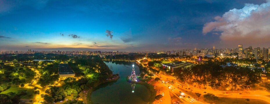 Aerial view of the illuminated Christmas tree in Ibirapuera Park, surrounded by lush greenery, a reflective lake, and the sprawling city skyline of Sao Paulo, Sao Paulo, Brazil.