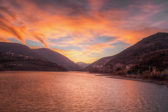 Aerial view of Lago di Caccamo at sunset with vibrant orange clouds reflecting on the water, surrounded by rolling hills and lush vegetation in Macerata, Marche, Italy.