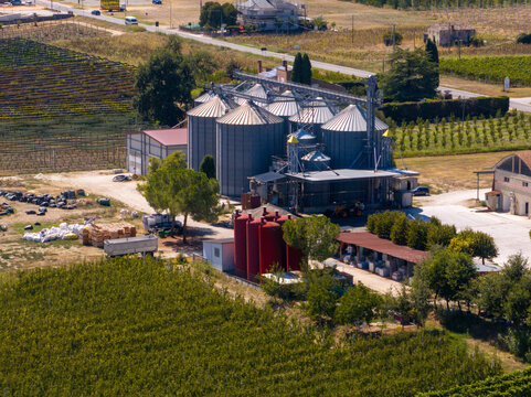 Aerial view of a grain silo facility with metallic storage bins and red tanks surrounded by agricultural fields and vineyards in Ortezzano, Marche, Italy.