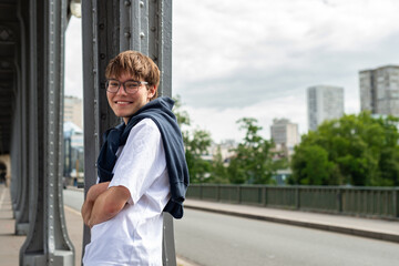 A smiling teenage boy wearing eyeglasses stands near a metal structure in Paris, France. He has a dark sweater tied around his neck and wears a white t-shirt outdoors. © dsbedarieva
