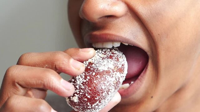 Close-up of a person's mouth taking a bite out of a sweet Indian dessert covered in coconut