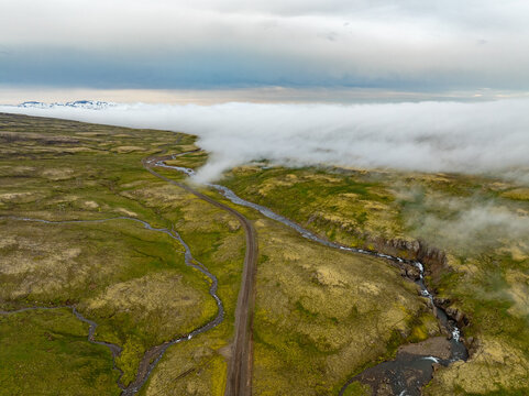 Aerial view of the Oxi mountain pass, featuring a winding road and stream through green mossy terrain with a thick layer of fog rolling over the hills in Mulathing, Iceland.