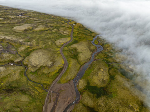 Aerial view of the winding road and river at Oxi, featuring vibrant green mossy terrain and a thick bank of fog rolling over the landscape, Mulathing, Iceland.