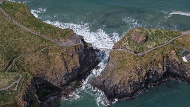Aerial view of the iconic Carrick-a-Rede rope bridge spanning the chasm between rugged cliffs, with waves crashing against rocks and a winding path, Ballintoy, Northern Ireland, United Kingdom.