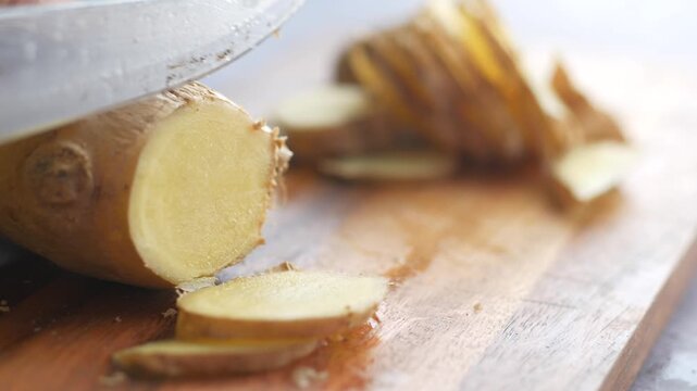 Close up of hand slicing fresh ginger root on wooden cutting board for cooking preparation