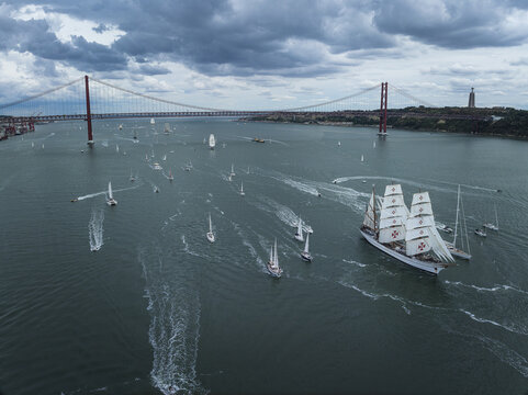 Lisbon, Portugal - 03 September 2026: Aerial view of a tall ship with white sails sailing on the Tagus River during a regatta, with the 25 de Abril Bridge in the background under a cloudy sky.