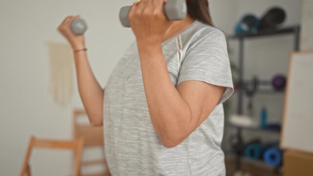 Woman lifting dumbbell with bent arm in studio fitness space, exercise ball and whiteboard visible; determination wellness.