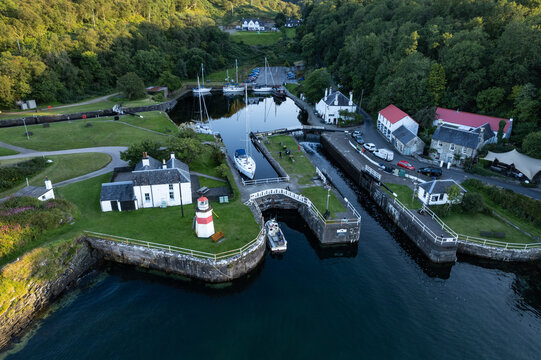 Lochgilphead, United Kingdom - 13 August 2022: Aerial view of Crinan Canal in Lochgilphead, Scotland, featuring a small lighthouse, moored boats in the lock, and surrounding lush green trees.