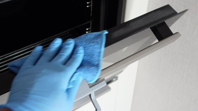 Person in blue gloves cleaning oven door glass with microfiber cloth during kitchen housekeeping routine