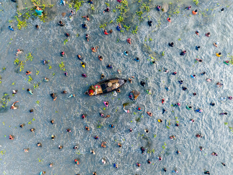 Pabna, Bangladesh - 28 November 2023: Aerial view of a small boat surrounded by many people wading in the water, with scattered aquatic plants and ripples on the surface.