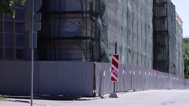 Urban scene shows building facade with scaffolding and protective netting; visible structural openings and weathered surfaces convey active renovation mood