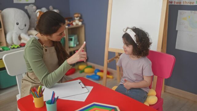 Woman teacher points finger at pink flashcard showing number one while child sits at red table in preschool building; early learning calm.