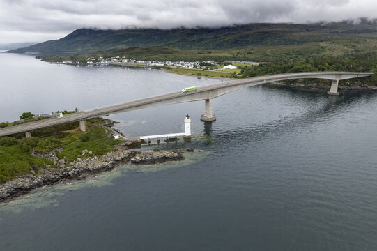 Isle of Skye, United Kingdom - 28 June 2021: Aerial view of the Skye Bridge spanning the water with a lighthouse below, surrounded by green hills, coastal scenery, and cloudy sky.