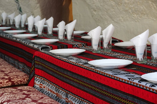 Close-up perspective of a communal banquet dining table setting inside a traditional underground troglodyte cave restaurant featuring patterned Amazigh tablecloths and glassware.