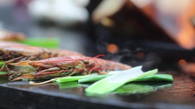 Fresh shrimp with green onion grilling on hot teppanyaki plate with smoke and flame. Seafood cooking process on iron griddle. Asian street food and restaurant cuisine concept.