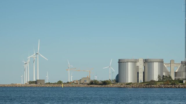 Wind turbines generate renewable energy along industrial coastal harbor in Copenhagen. Modern facility with storage silos supports green electrical power generation for sustainable future.