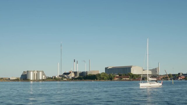 White sailboat cruises past an industrial power plant in Copenhagen harbor. Nautical yacht navigates calm sea water on a sunny day. Coastal maritime transit supports active vacation travel.