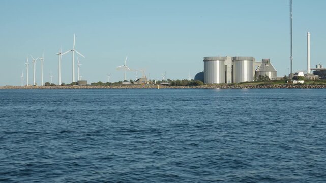 Wind turbines rotate on a rocky breakwater along an industrial coastline in Copenhagen. Offshore wind farm generates clean electricity. Sustainable energy facility supports eco friendly power.
