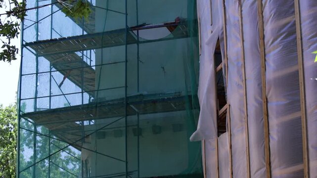 Urban scene shows building facade with scaffolding and protective netting; visible structural openings and weathered surfaces convey active renovation mood