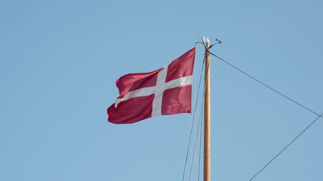 National flag of Denmark flutters on a wooden mast against a clear blue sky. Red Dannebrog banner waves in the wind. This patriotic Danish symbol represents Scandinavian culture and pride.