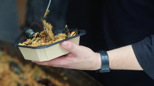 Man holding takeout box with fried rice, whole prawn and mussels with green sauce. Street food meal in plastic container. Seafood take away dish concept.