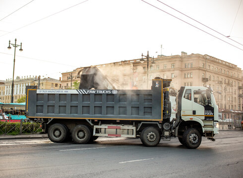 Heavy Chinese FAW Trucks J6 dump truck tipper during asphalt road construction works. Industrial vehicle with a large tipper body unloads hot smoking asphalt mix material on a city street background.