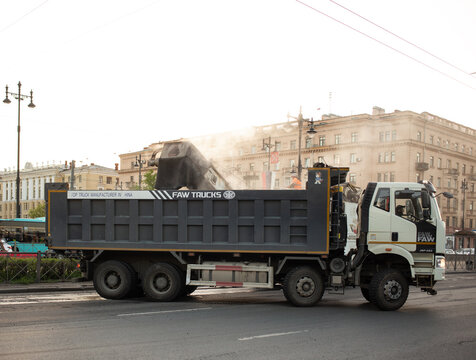 Heavy Chinese FAW Trucks J6 dump truck tipper during asphalt road construction works. Industrial vehicle with a large tipper body unloads hot smoking asphalt mix material on a city street background.