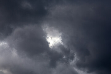 Fototapeta premium Dramatic dark storm clouds filling the sky with a small bright white sun light break. Overcast weather scene before a heavy rain. Natural atmospheric backdrop and texture template for graphic design. 