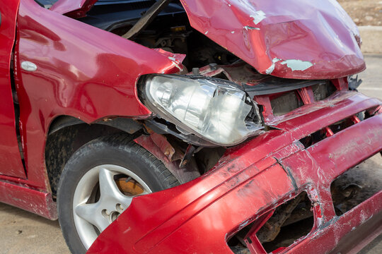 Damaged red sport compact car after a severe traffic accident on a city street side. Smashed front bumper, crumpled bonnet hood, broken headlight optics, and crushed fender panel. Auto insurance. 