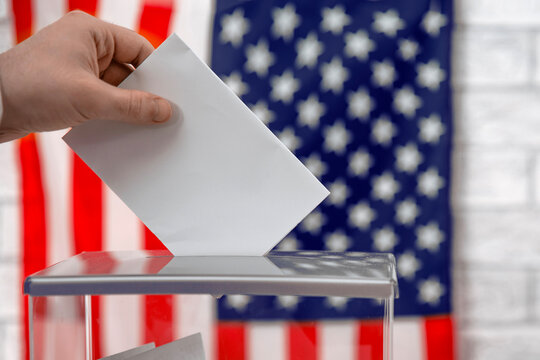 Voter casting a voting ballot into a transparent ballot box with the US flag. American elections, democracy and voting rights.