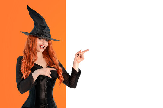 Halloween Witch, red-haired woman in witch costume and hat pointing to empty board for copy space. Halloween concept, spooky holiday mockup for text or advertising design.