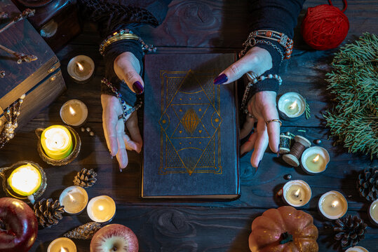 Witch's hands witchcrafting with a wizard's black book, casts a spell among candles, pumpkins on Halloween night