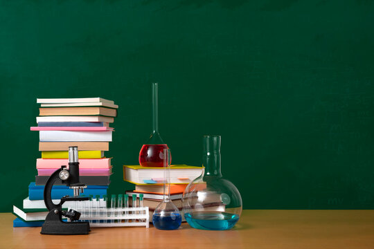 School background, chemistry class concept. Microscope, textbooks and chemistry glassware with colored solution in front of chalkboard. Empty space, copy space.