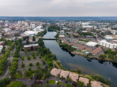 Spokane, United States - 15 August 2026: Aerial view of the Spokane River flowing through the city with downtown buildings, bridges, and lush greenery under a hazy sky.