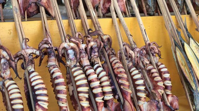 Detailed close-up of smoked squid on bamboo skewers at a local market, with a customer hand briefly pointing at the products.
