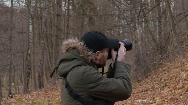 Standing photographer raising camera to eye on leaf-strewn autumn forest trail with bare trees and dense trunks in olive parka with fur-trimmed hood under overcast sky