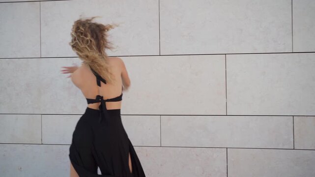 Beautiful young woman dancing gracefully on the street. Elegant dancer in black dress against city wall on sunny day. Seductive dancer with curly blonde hair 