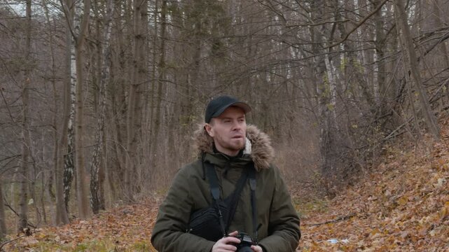Rear view of photographer in olive parka with fur-trimmed hood standing on autumn trail, holding camera to eye among leaf litter, bare deciduous branches, and vertical trunks under gray overcast light